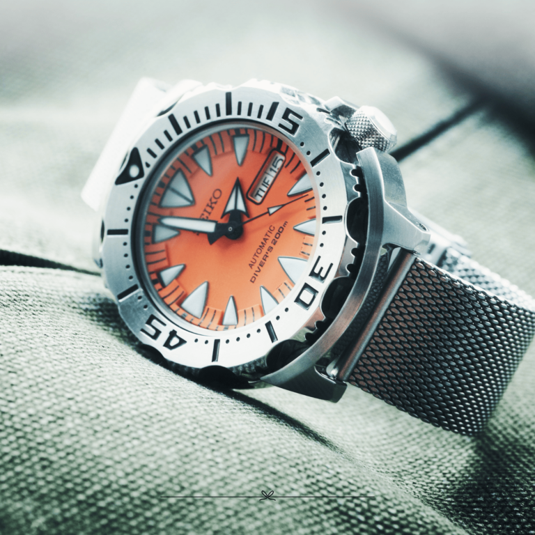 Silver wristwatch with an orange face and black markings, set on a textured green fabric background.