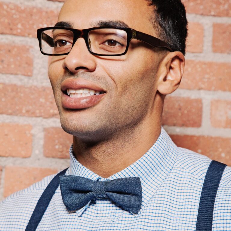 A man sporting glasses and showcasing one of the finest bow ties for men stands confidently against a brick wall.