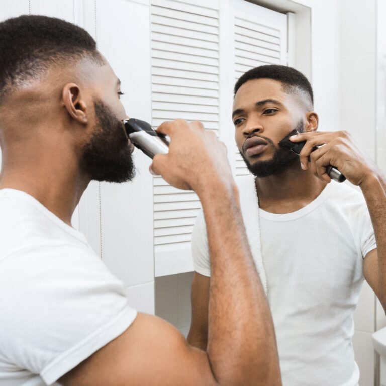 A man trims his beard with an electric trimmer while looking in the bathroom mirror.