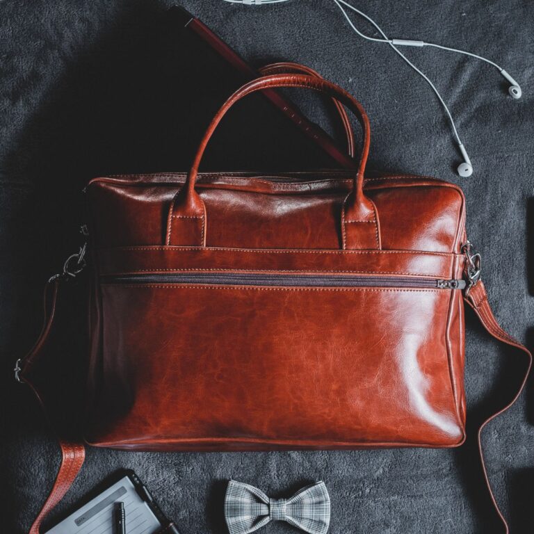 A brown leather men's bag rests on a dark surface, accompanied by earphones, a pen, a notepad, and a bow tie nearby.