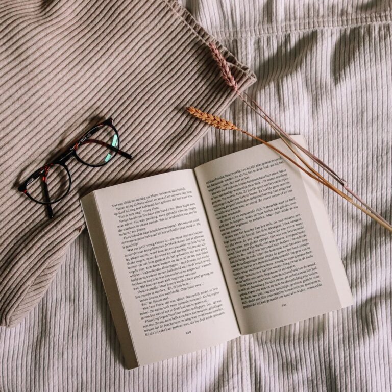 An open book rests on a beige blanket with a pair of eyeglasses and two dried wheat stalks beside it.