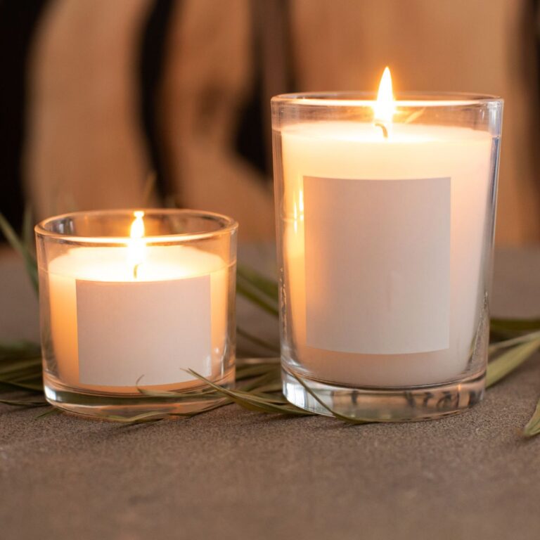 Two lit candles in clear glass holders on a gray surface with scattered dried leaves around them.