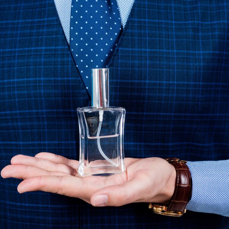 A person in a blue checkered suit holds a clear glass cologne bottle in their palm.