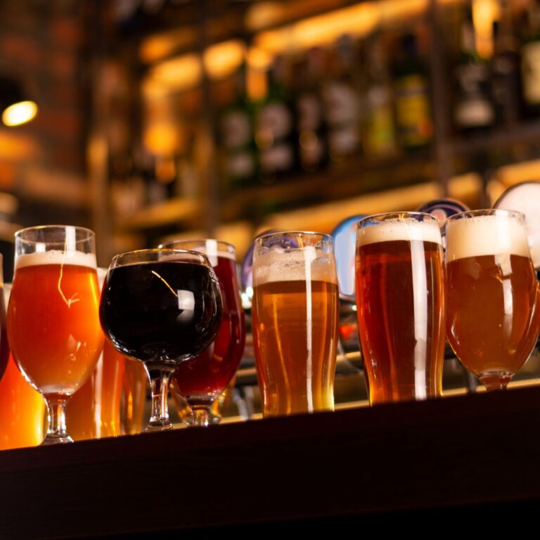 A row of various beer glasses, filled with different types of beer, sits on a bar counter with a blurred background of bottles.