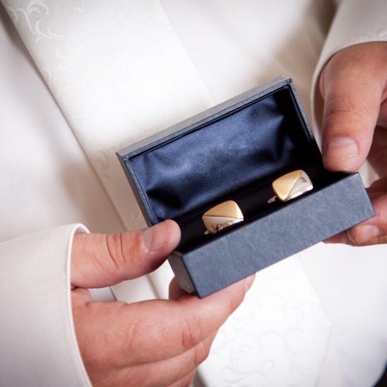 A person in a white suit holds an open box containing gold cufflinks.