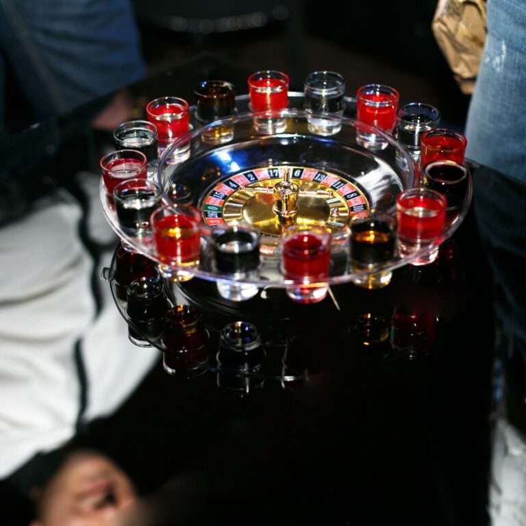 A roulette wheel surrounded by shot glasses filled with various colored liquids on a reflective surface.