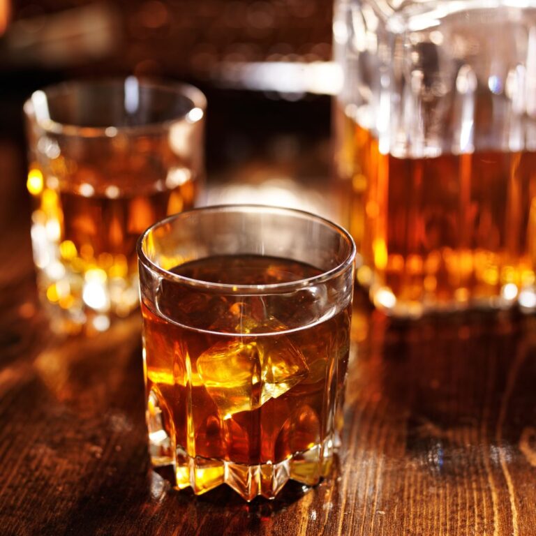 Three glasses of amber liquid, likely whiskey, are placed on a wooden table. The foreground glass is partially filled with ice cubes.