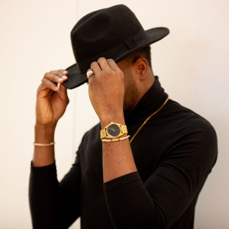 A stylish individual in a sleek black outfit adjusts one of the latest mens hats, showcasing a gold watch and rings that perfectly complement the ensemble.