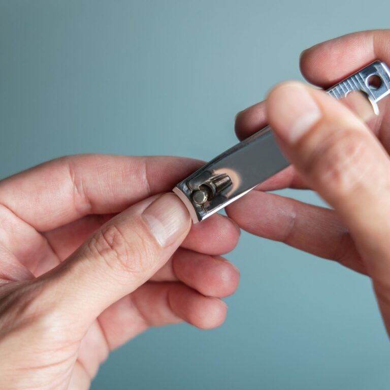 A person trims their nails with a metal nail clipper against a blue-grey background.