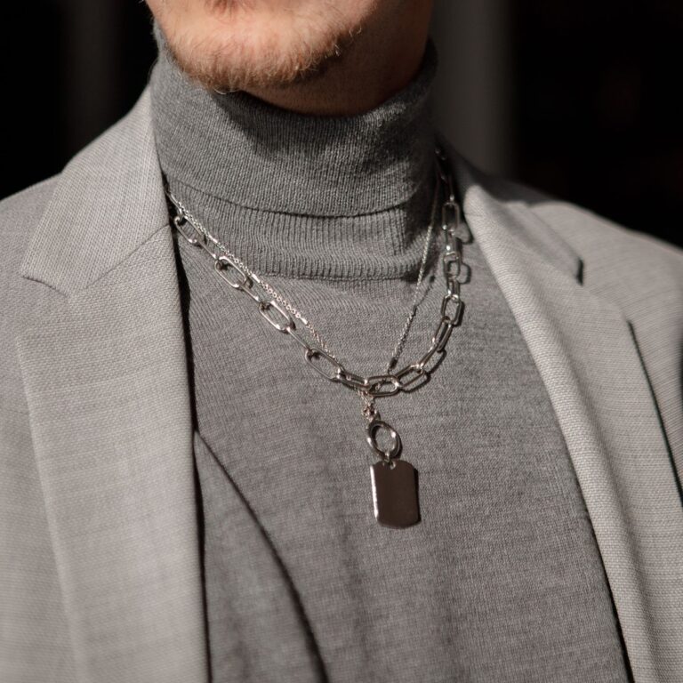 Person wearing a gray turtleneck, a light gray blazer, and two silver necklaces with a rectangular pendant.