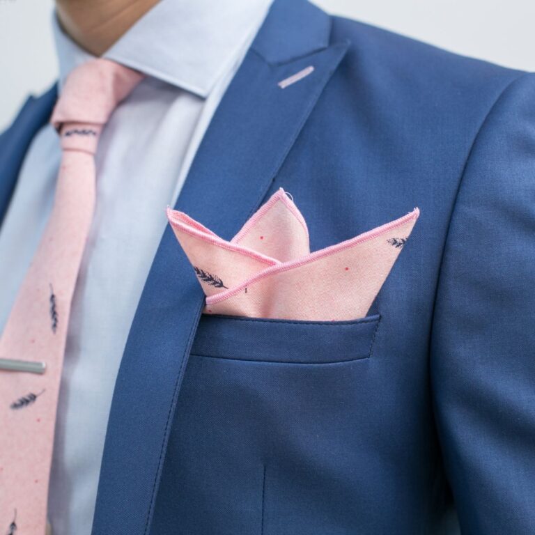 Close-up of a person wearing a blue suit with a pink tie and a neatly folded pink pocket square.