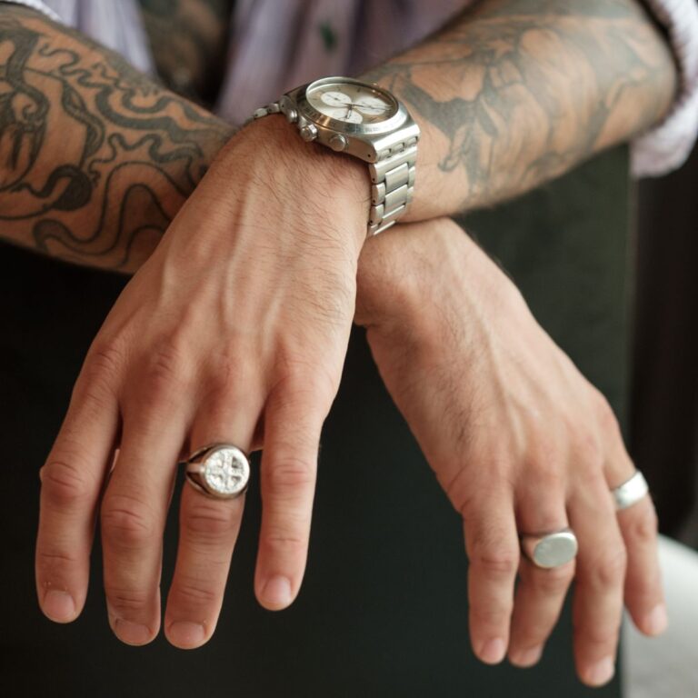 Close-up of tattooed arms crossed at the wrists, wearing a wristwatch and several rings.