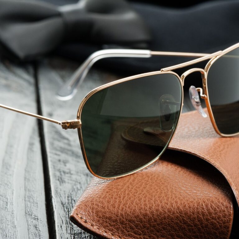 A pair of sunglasses with a golden frame rests on a brown leather case, alongside a black bow tie, on a wooden surface.