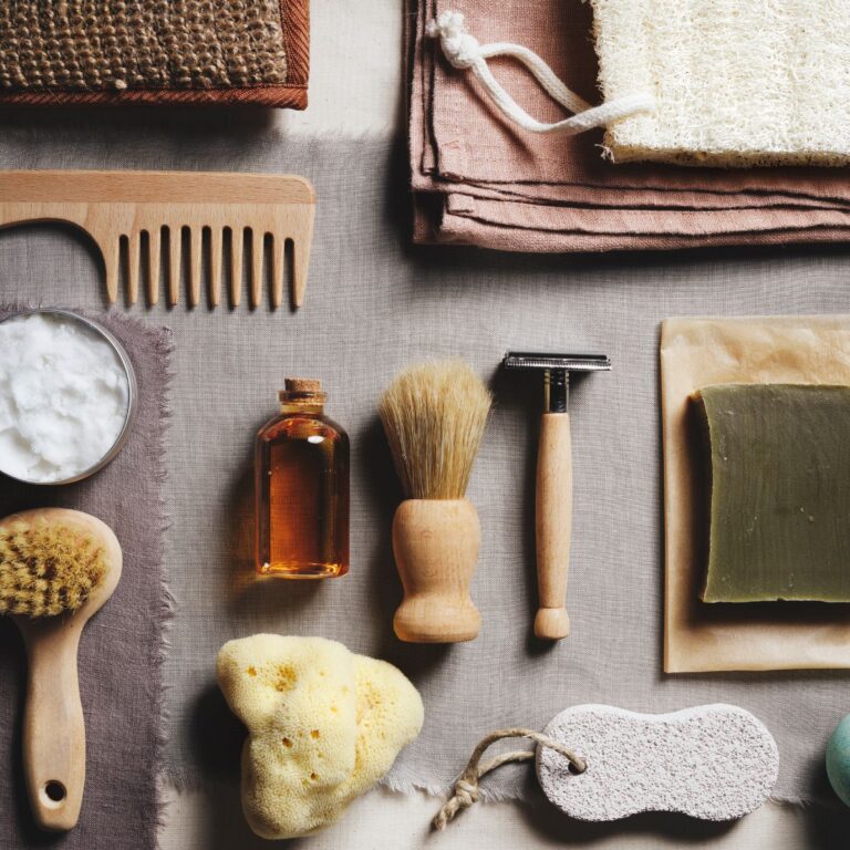 A collection of grooming items including a wooden comb, brush, shaving brush, razor, bar soap, and natural sponge laid out on fabric.