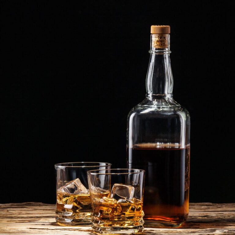 A bottle of whiskey, perfect as a whisky gift for men, sits next to two glasses filled with whiskey and ice cubes, elegantly arranged on a wooden surface against a dark background.