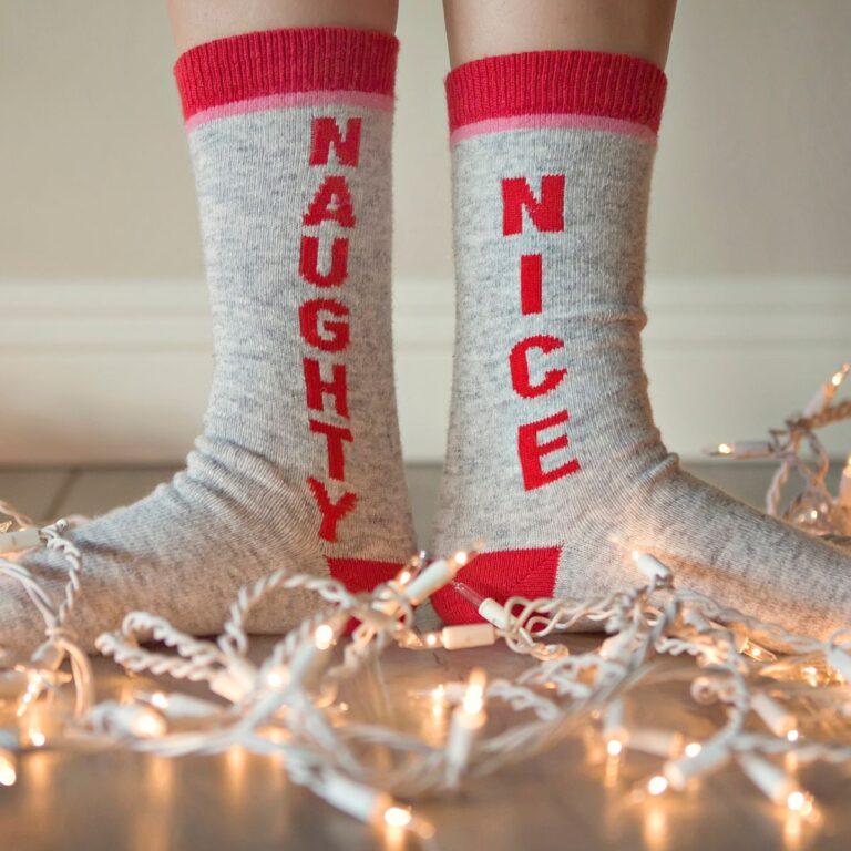 Two gray socks with red cuffs. One sock says "NAUGHTY," the other says "NICE." They are surrounded by string lights on the floor.