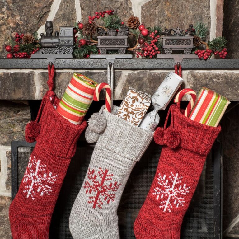 Three Christmas stockings filled with candy canes and gifts hang from a fireplace mantel decorated with a toy train, pine cones, and berries.