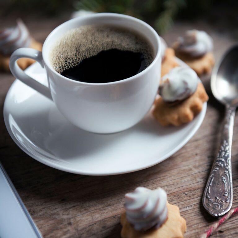 A cup of black coffee, perfect for coffee lovers, sits on a saucer surrounded by small cookies, with a spoon resting nearby on the wooden table.