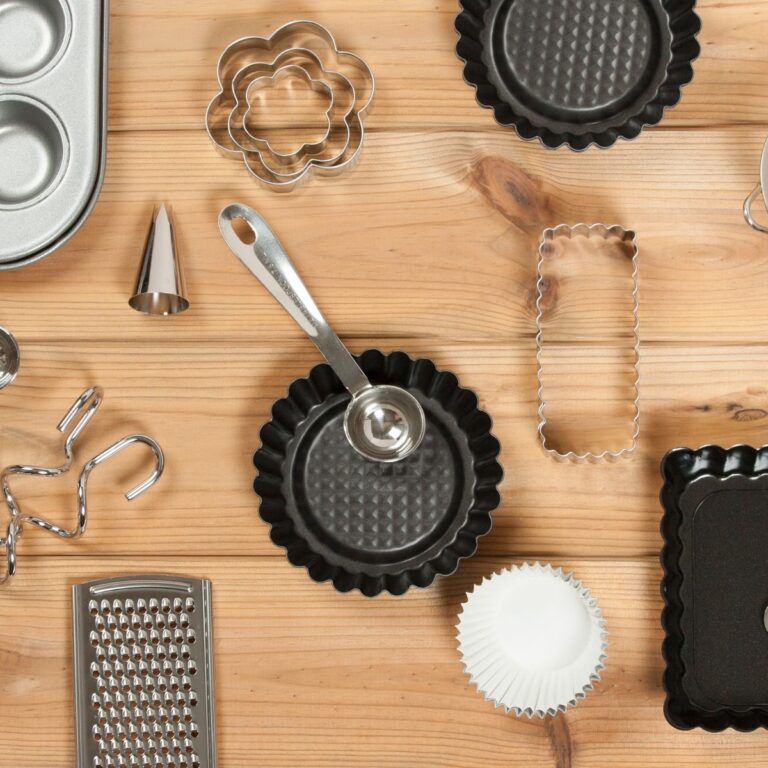 Baking tools on a wooden surface, including muffin tray, tart pans, cookie cutters, measuring spoon, and a grater.