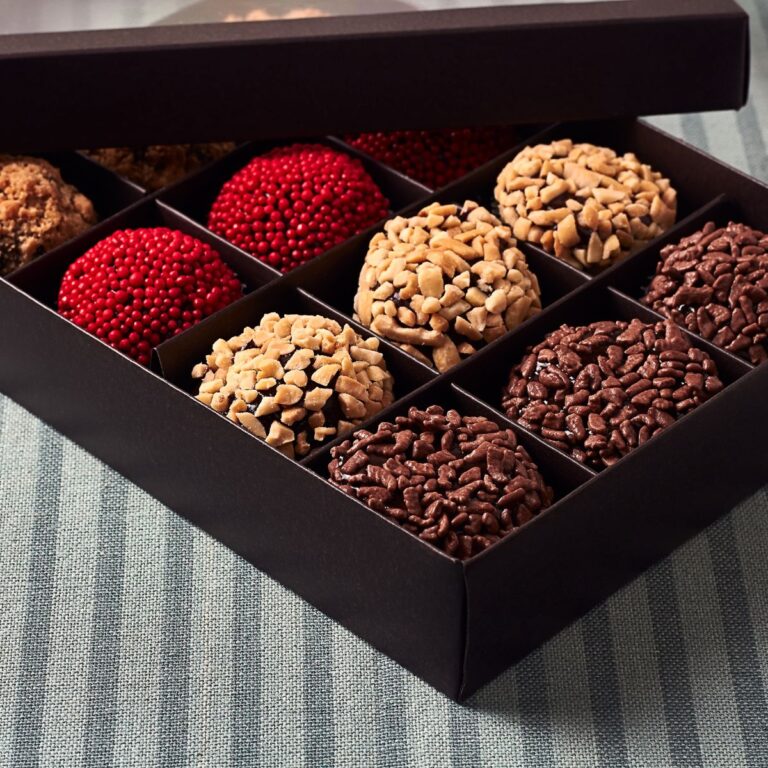 A black box containing gourmet treats of assorted chocolate truffles, including ones coated with nuts, red sprinkles, and chocolate sprinkles, rests elegantly on a striped surface.