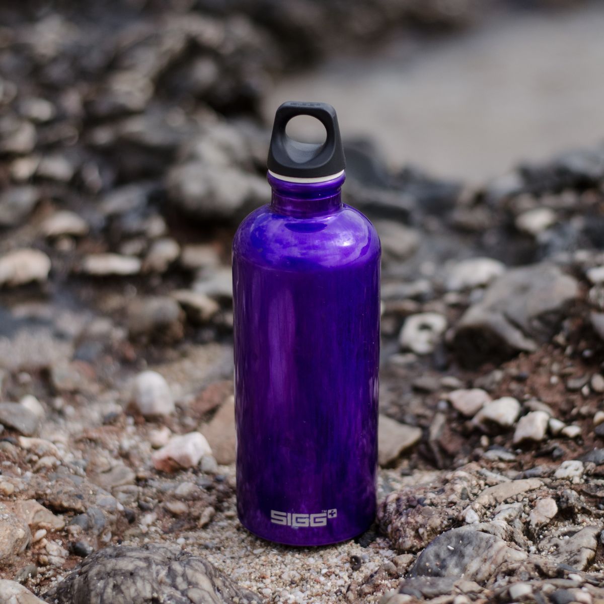 A purple SIGG, the epitome of an environmentally friendly water bottle, with a sleek black cap, stands proudly on rocky terrain outdoors.