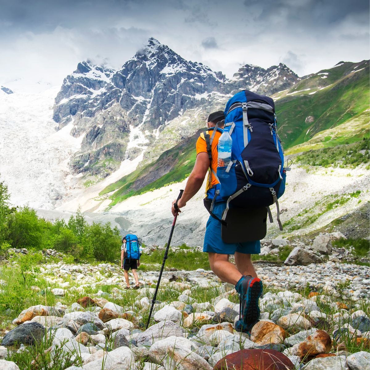 Two hikers with hiking backpacks and trekking poles navigate a rocky trail towards snow-capped mountains, enveloped by lush greenery under overcast skies.